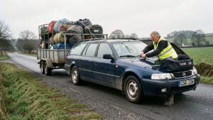 Un homme vérifie les sangles d’une remorque chargée attelée à une voiture sur une route de campagne.